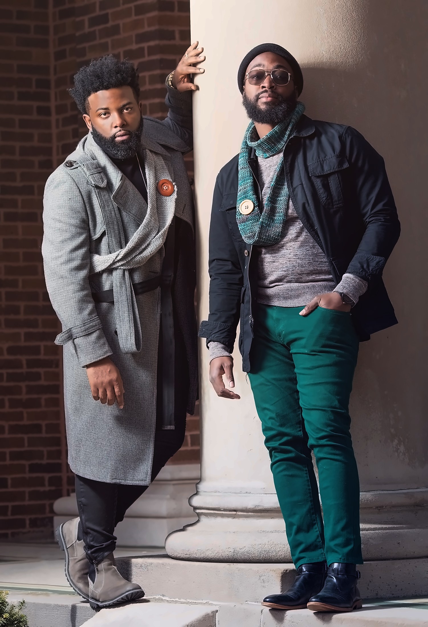 Two friends in stylish coats posing against a brick column