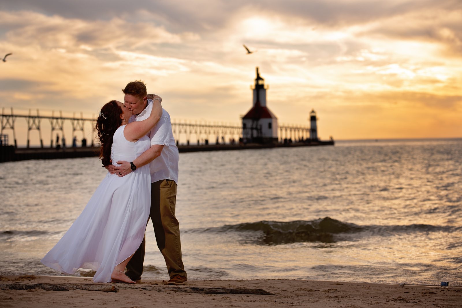 Bride and groom embracing beside the lighthouse on a Lake Michigan beach at sunset