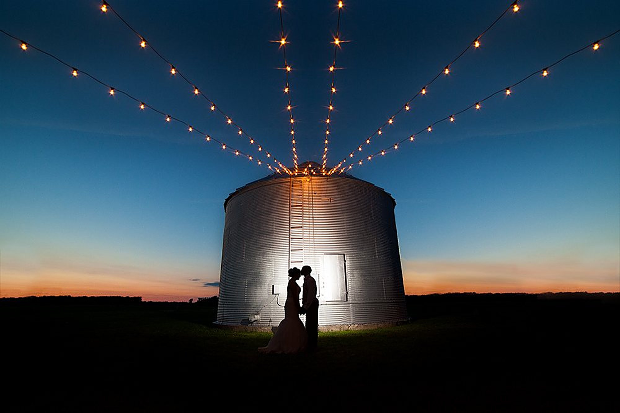 Bride and groom silhouetted under string lights beside a grain silo at dusk