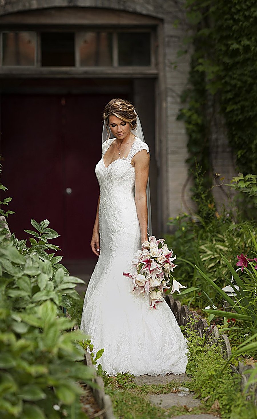 Bride in a lace gown holding a lily bouquet outside a garden doorway