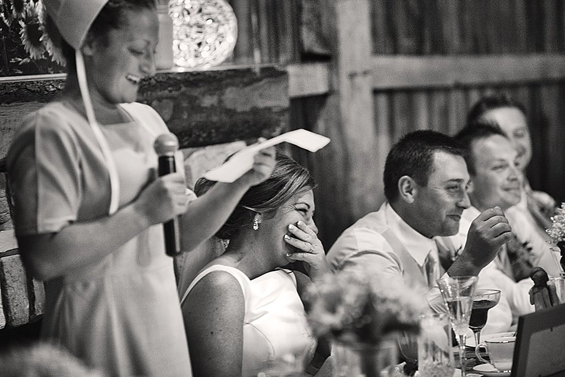 Black-and-white candid of a maid of honor giving a toast as the bride laughs through happy tears